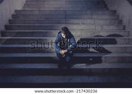 Young desperate man in casual clothes abandoned lost in depression sitting on ground street concrete stairs alone suffering emotional pain, sadness, looking sick in grunge lighting