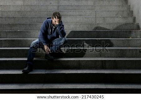Young desperate man in casual clothes abandoned lost in depression sitting on ground street concrete stairs suffering emotional pain, sadness, looking sick in grunge lighting