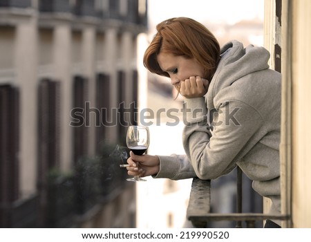 young attractive woman suffering depression and stress smoking drinking glass of wine at the balcony window in pain and grief feeling sad and desperate in urban background