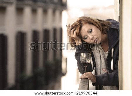 young attractive woman suffering depression and stress smoking drinking glass of wine at the balcony window in pain and grief feeling sad and desperate in urban background