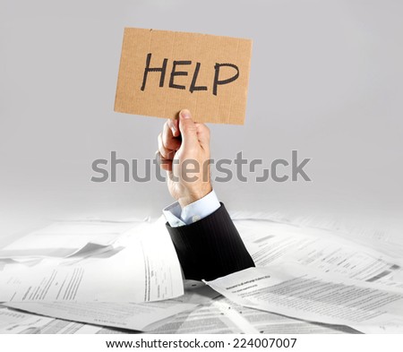 Hand of caucasian businessman emerging from office desk loaded of paperwork , invoices and a lot of papers and documents holding message card  asking for help