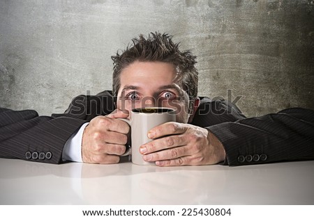 young addict business man in suit holding cup of coffee anxious and crazy in maniac caffeine addiction needing to keep awake leaning on desk on grunge studio background