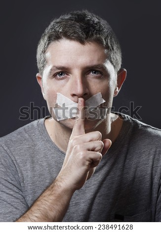 attractive young man with mouth sealed on duct tape to prevent him from speaking keeping him mute and censored in freedom of speech and expression concept isolated on dark grunge studio light style