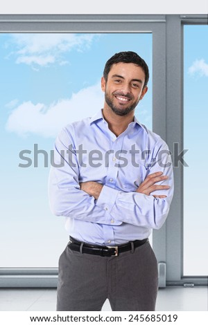 young attractive businessman smiling happy standing in corporate portrait smiling with folded arms in smart casual shirt and suit trousers in front of blue sky office window view in success concept