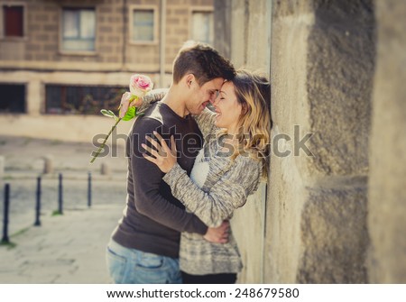 candid portrait of beautiful European couple with rose in love kissing on street alley celebrating Valentines day with passion against stone wall on urban background