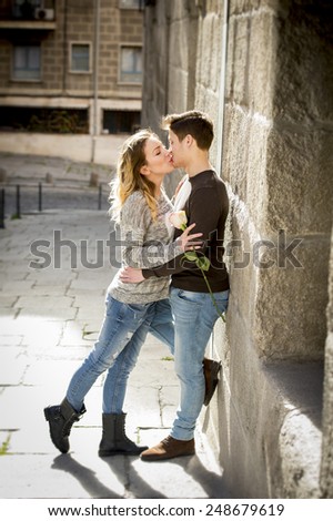 candid portrait of beautiful European couple with rose in love kissing on street alley celebrating Valentines day with passion against stone wall on urban background