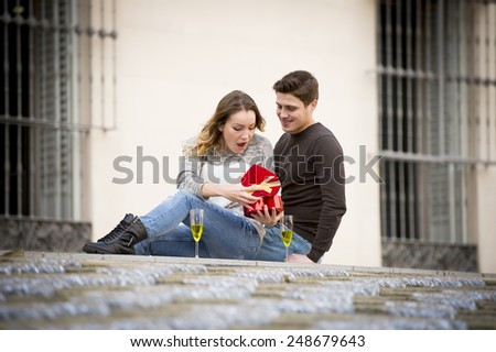 couple in love kissing on street celebrating Valentines day in Champagne toast with girl receiving heart shaped box present in urban background