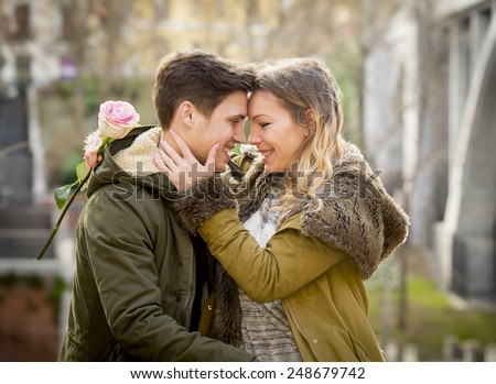 candid portrait of beautiful European couple with rose in love kissing on street  celebrating Valentines day with passion sitting on city park in winter urban background