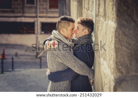 young happy attractive gay men couple holding rose hugging and kissing each other outdoors on street in free homosexual love concept in Valentines day on urban background