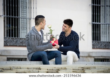 young cool gay men couple on street with rose flower and heart shape box present giving gift celebrating valentines day in love on street urban background