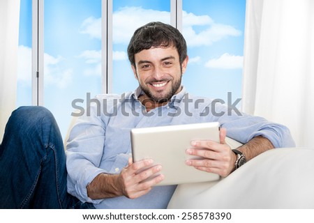 Young attractive Hispanic man at home sitting on white couch using digital tablet or pad looking relaxed smiling at living room enjoying surfing internet watching online movie