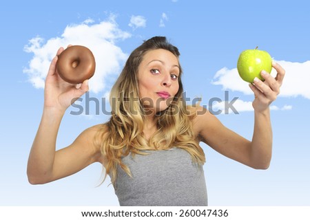 young attractive sport woman holding apple and chocolate donut in her hands in healthy fruit versus sweet junk food dilemma in fitness, body health care and healthy nutrition concept