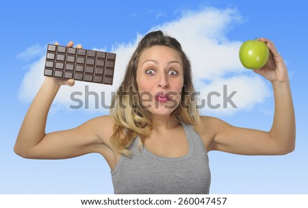young attractive sport woman holding apple and chocolate bar in her hands in healthy fruit versus sweet junk food temptation in fitness, body health care and healthy nutrition concept