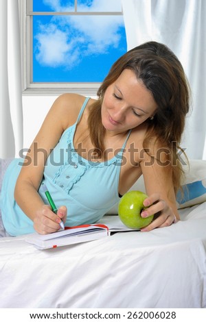 young attractive and beautiful woman in nightgown lying on bed at bedroom holding apple writing diary with pen in relax looking tranquil and happy in American lifestyle concept