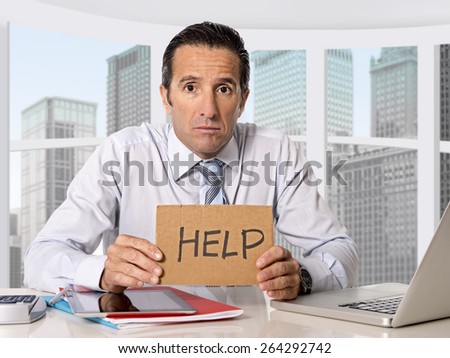 desperate senior businessman in crisis working on computer laptop at business district office desk in stress under pressure facing work problems asking for help holding sign billboard