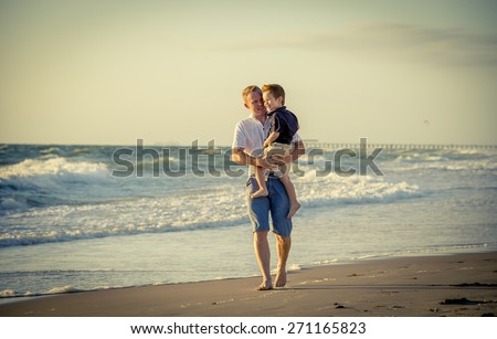 young happy father holding in his arms little son walking on the beach with barefoot in sand in front of sea waves, the kid smiling and having fun together with dad in Summer vacation concept