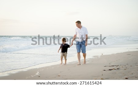 young happy father holding holding hand of little son walking together on the beach with barefoot in sand in front of sea waves, the kid smiling and having fun with dad in Summer coast holidays