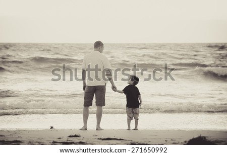 young happy father holding hand of little son walking together on the beach with barefoot in sand in front of sea waves, the kid smiling and having fun  with dad in Summer sunset coast
