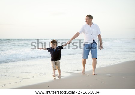 young happy father holding hand of little son walking together on the beach with barefoot in sand in front of sea waves, the kid smiling and having fun with dad in Summer coast holidays
