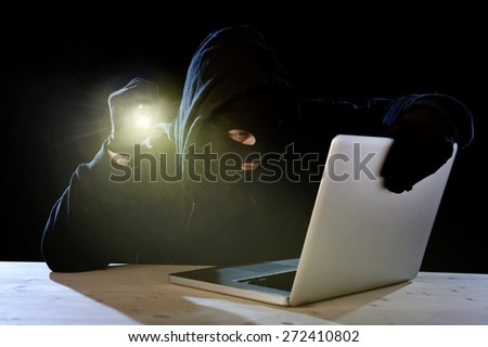 hacker man in black hood mask with computer laptop holding flashlight  in dangerous dark look hacking system having access to data info and privacy in business digital crack and cyber crime concept