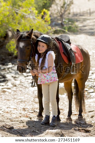 sweet beautiful young girl 7 or 8 years old holding bridle of little pony horse smiling happy wearing safety jockey helmet posing outdoors on countryside in summer holiday
