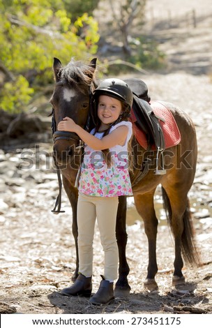sweet beautiful young girl 7 or 8 years old hugging head of little pony horse smiling happy wearing safety jockey helmet posing outdoors on countryside in summer holiday