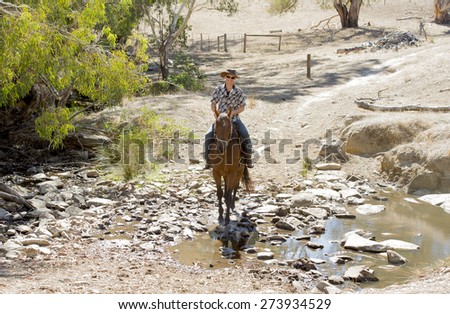 young horse instructor or cattleman riding the animal wearing sunglasses, cowboy hat and rider boots looking cool while taking a ride at countryside summer landscape