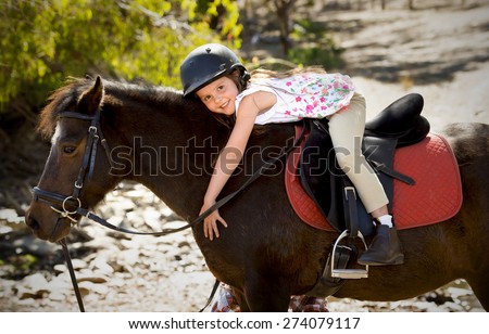 sweet beautiful young girl 7 or 8 years old riding pony horse hugging and smiling happy wearing safety jockey helmet posing outdoors on countryside in summer holiday