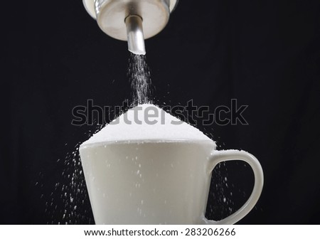 man hand with sugar bowl pouring a crazy lot of it spilling out everywhere in full coffee cup in insane sugar addiction and unhealthy nutrition concept isolated on black background