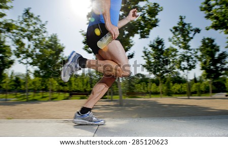 close up athletic legs of young man holding isotonic energy drink while running in city park with trees on the background on summer training session fitness and healthy lifestyle concept