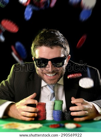 happy young attractive man grabbing and protecting poker chips with his hands after winning bet gambling at Casino with chips flying all around isolated on black background
