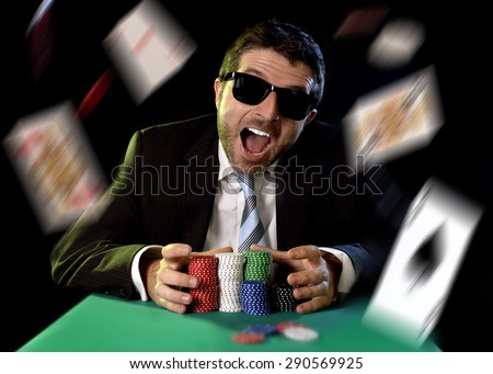 happy young attractive man grabbing poker chips after winning bet gambling on table with playing cards on green felt at Casino and chips and cards flying all around