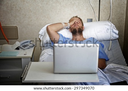 young man in hospital room lying in bed sick and injured using internet in computer laptop feeling sad and depressed researching info on his own disease injury or sickness
