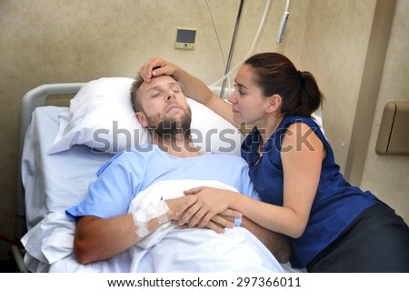 young sick man lying in bed at hospital room after suffering accident having his worried and caring wife or girlfriend together holding his hand giving him love and support