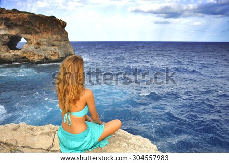 young attractive woman looking at sea horizon sitting alone on rock cliff in cyan glamour dress under a summer blue sky in relax , meditation and serenity concept