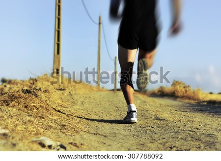 back view of young sport man legs and feet running on countryside track with power line poles training in summer harsh light in cross country runner concept and healthy fitness lifestyle