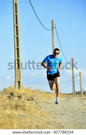 frontal view of young sport man with sun glasses running on countryside track with power line poles training in summer in cross country runner concept and healthy fitness lifestyle