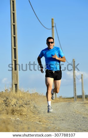 frontal view of young sport man with sun glasses running on countryside track with power line poles training in summer in cross country runner concept and healthy fitness lifestyle