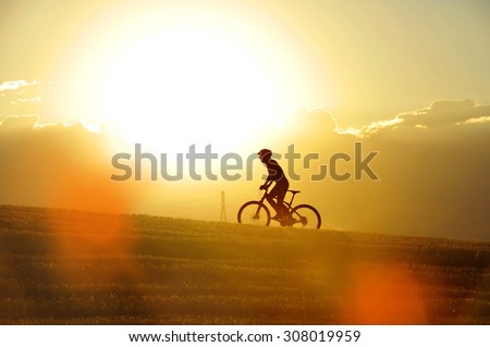 profile silhouette of sport man cycling uphill riding cross country mountain bike on sunset field with harsh sun light and high contrast in amazing beautiful rural landscape with orange lens flare