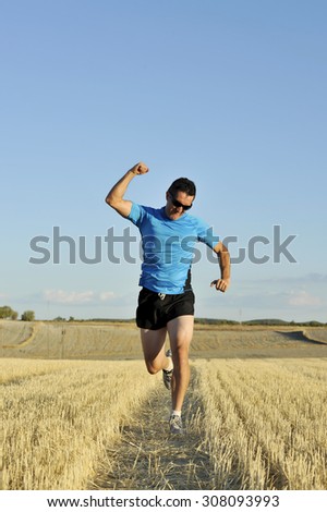young sport man with sunglasses running outdoors on straw field ground in frontal perspective towards camera doing victory sign with fist in healthy lifestyle and competition concept