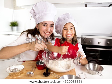 happy mother baking with little daughter in apron and cook hat working with flour , bowl and spoon preparing dough teaching the kid baking and having fun together