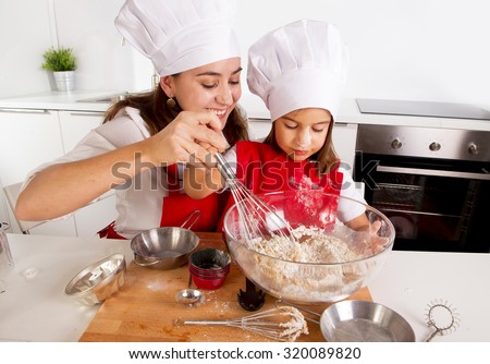 happy mother baking with little daughter in apron and cook hat working with flour , bowl and spoon preparing dough teaching the kid cooking and having fun together