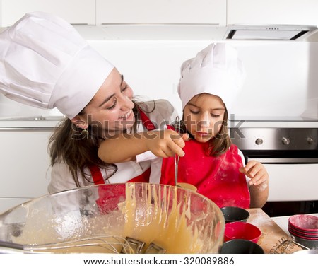 happy mother baking with little daughter in apron and cook hat filling mold muffins with dough and chocolate teaching the kid cooking and having fun together