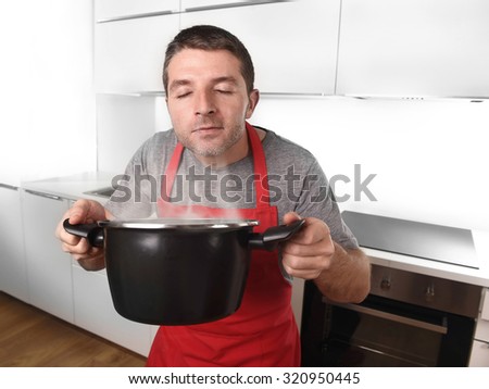 young man at home kitchen in red cook apron holding pot delighted with the good smell of his dish enjoying the delicious aroma in delightful face expression