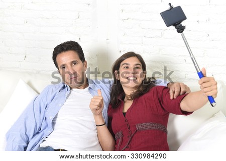 young attractive couple with woman taking selfie photo or shooting self video with mobile phone and stick and man bored tired and sick of pictures sitting at home  in technology image concept