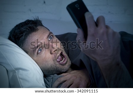 young man in bed couch at home late at night with intense face expression using mobile phone in low light watching online porn enjoying alone in internet addiction