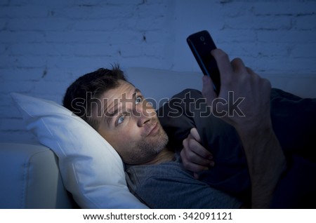 young man in bed couch at home late at night with intense face expression using mobile phone in low light watching online porn enjoying alone in internet addiction