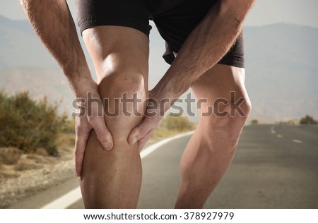young sport man with strong athletic legs holding knee with his hands in pain after suffering muscle injury during a running workout training in asphalt road in muscular or ligament wound