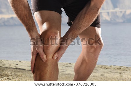 young sport man with strong athletic legs holding knee with his hands in pain after suffering muscle injury during a running workout beach training in muscular or ligament wound