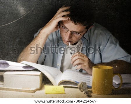 Young student at home desk reading biting pen studying at night with pile of books and coffee cup preparing exam in university education concept in edgy  light set